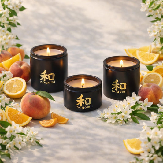 Lit Nagomi Citrus Vitality candles in black glass jars on a sandy surface with orange slices, peaches, and jasmine flowers