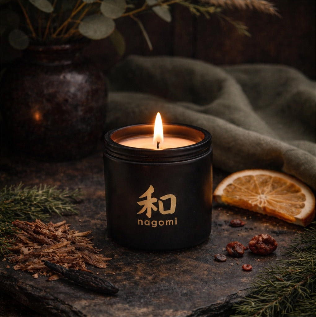 Lit 8 oz Nagomi Dragon’s Whisper candle in a black glass jar outdoors with oranges, cedarwood shavings, and tonka beans