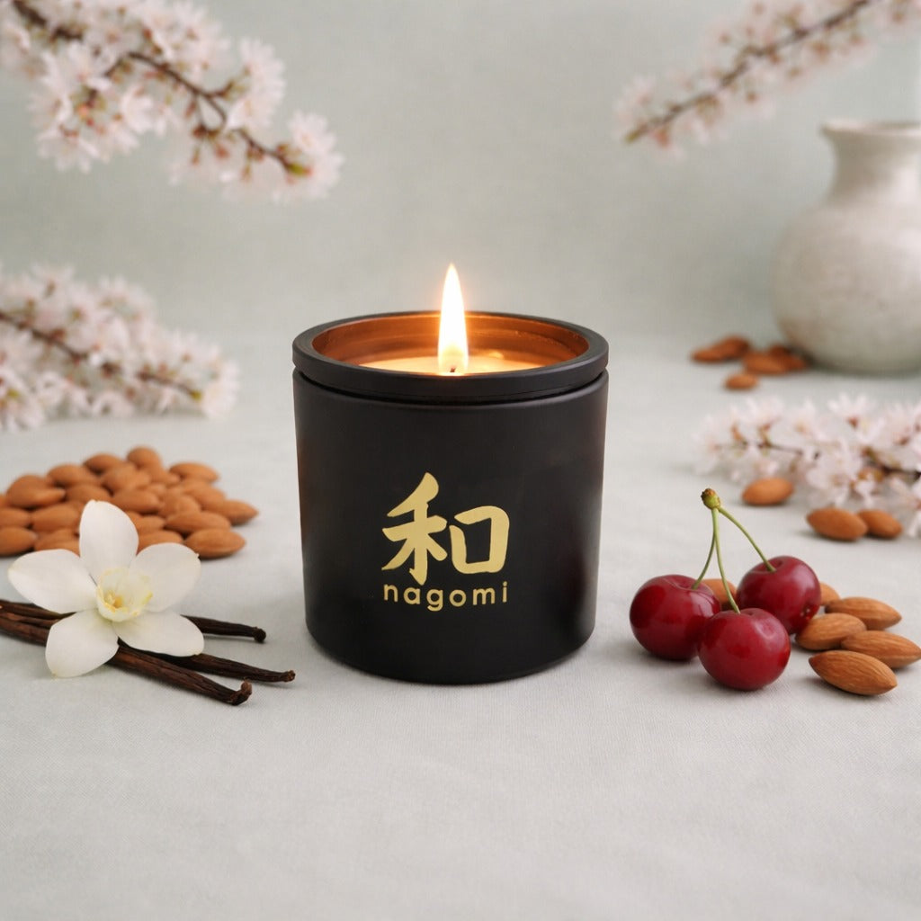 An eight-ounce Nagomi Aromatic Journey candle in a black glass jar, lit, on a neutral background with cherries, almonds, and vanilla beans.