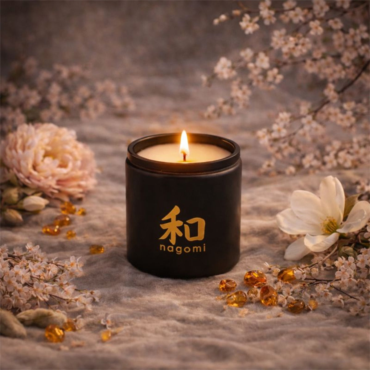 Lit 8oz Nagomi Blossom Odori candle in a black glass jar on sandysurface with cherry blossoms, magnolia flowers, and amber