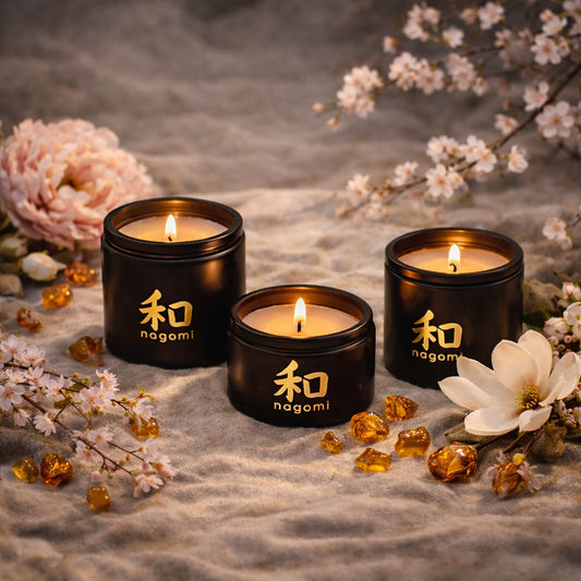 Lit Nagomi Blossom Odori candles in black glass jars on a sandy surface with cherry blossoms, magnolia flowers, and amber pieces