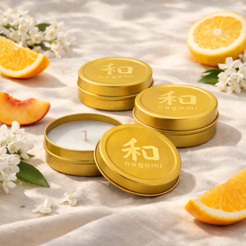 Lit Nagomi Citrus Vitality mini-tin candles arranged on sand with orange slices, peaches, and jasmine flowers