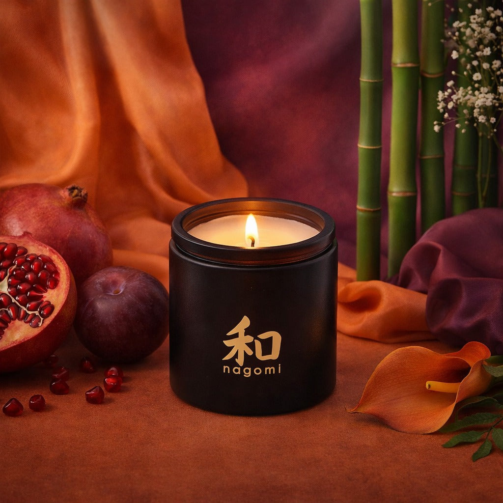 Twelve-ounce Nagomi Eternal Ascent Candles in a  black glass jar, lit, on a neutral background with oranges, plums and bamboo