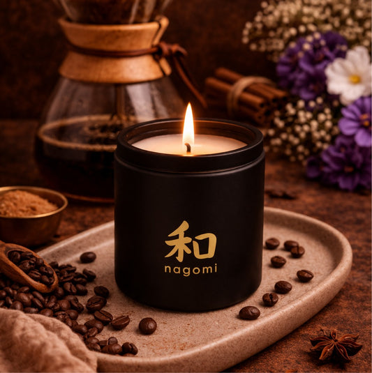 An eight-ounce Nagomi Mindful Brew candle in a black glass jar, lit, on a neutral background with fruit, coffee, and african violets